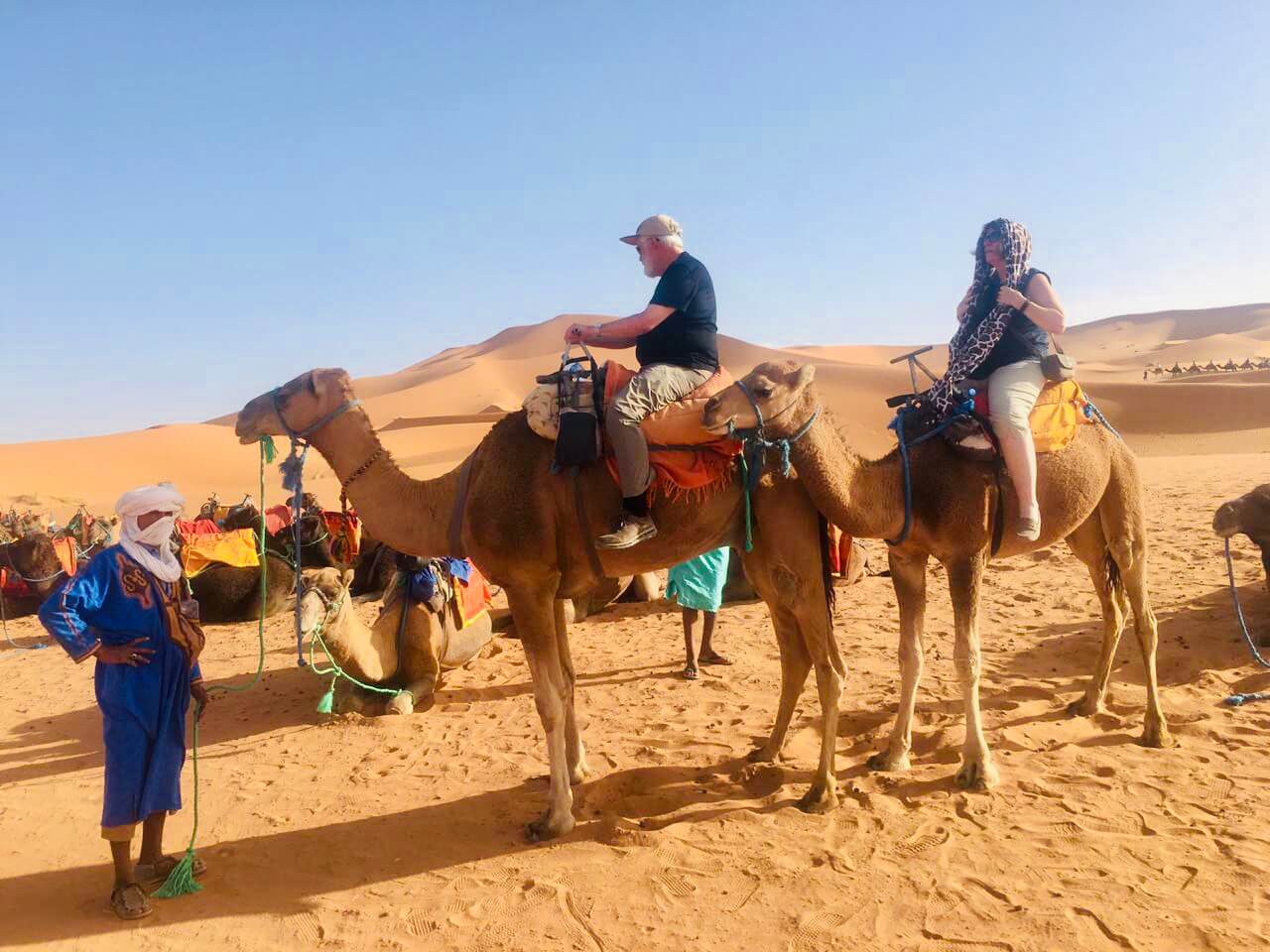 Two older Gateway2Morocco travellers riding camels into the Erg Chebbi dunes at Merzouga, with their Berber camel handler leading on foot